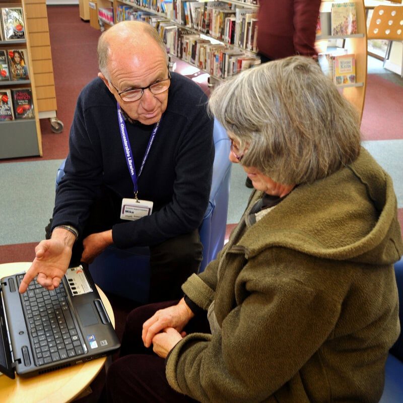 A local library volunteer helps a woman with an IT question.